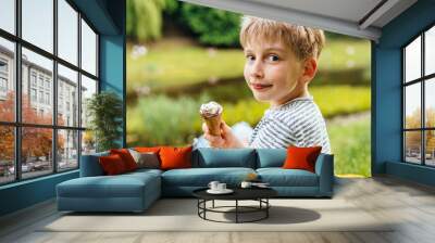 Cute school age blond white boy sitting on lake side, turn back, looking at camera, relaxing, eating ice cream at summer day outdoor. Vacation, leisure, lifestyle and childhood concept. Wall mural