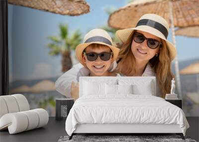 Close up portrait of young woman in hat and sunglasses embracing her son. Mother and kid enjoying summer vacation in tropical country with palm trees on background. Wall mural