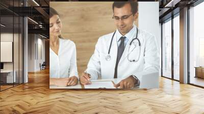 Doctor and patient discussing medical exam results while sitting at the desk in clinic. Male physician using clipboard for filling up medication history record of young woman. Data in medicine Wall mural