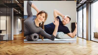 Gorgeous woman instructor performing back exercise on a foam roller at same time with handsome guy at pilates studio. Coach and patient doing fascia exercise on side surface of back. Wall mural