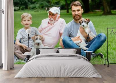 Grandfather, father and son sits, hugs, caress their two dogs Jack russel terrier on green grass in the summer park. Family Vacation. Generational connection. Pet, love friendship and people concept Wall mural