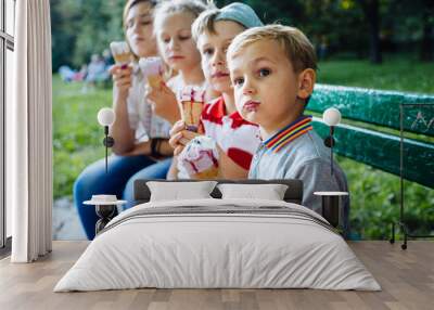 Group of four happy children eating ice cream together outdoor. Photo of happy blond girls with two handsome boys sitting on the bench and smiling at camera. Wall mural