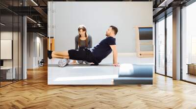 Handsome man using a foam roller on the ground, female rehabilitation specialist instructor help him in modern pilates studio. Rehabilitation after trauma concept. Wall mural