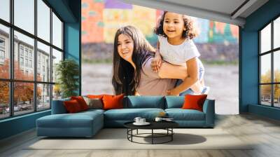 Happy family piggyback. Parenthood, childhood, love and happiness concept. Single mixed race mother playing with her cute curly preschooler daughter outdoor in city street, bright blurred background. Wall mural