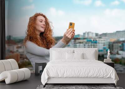 happy young lady with red curly hair exploring city and taking selfies on mobile phone Wall mural