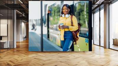 Lifestyle photo of young black woman come out while opening door to tram. Portrait of female traveler In yellow blouse with backpack smiling drinking coffee on bus stop. Wall mural