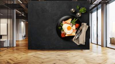 Overhead shot of breakfast or lunch with fried egg, bread toast, green asparagus, tomatoes and bacon on black plate with copy space. Wall mural