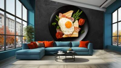 Overhead shot of breakfast or lunch with fried egg, bread toast, green asparagus, tomatoes and bacon on black plate with copy space. Wall mural