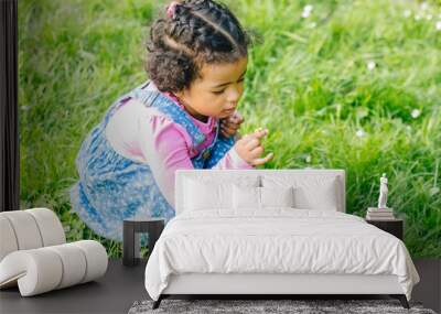 Outdoor close up portraot of hispanic toddler girl three years old in blue denim sarafan and pink shirt picking and smelling to flowers, sit at grass at summer park. Wall mural