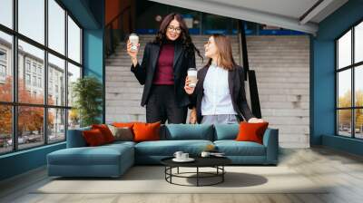 Outdoors fashion portrait of two cheerful girls drinking coffee. Walking in the city, moving down stairs. Wall mural