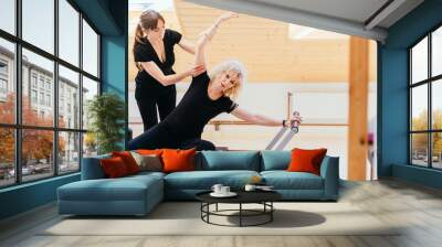 Pilates lesson on reformer, personal coach in black sportswear workout with sporty eldery beautiful blond woman. Rehabilitation and healty lifestyle concept. Wall mural