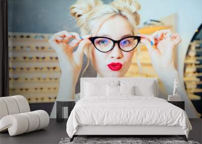 Portrait of fashion woman with red lips and funny hairstyle wearing and buying sunglasses with optician modern shop interior behind her. Close up. Wall mural