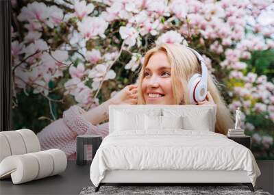 Teenager hipster inspiring girl in headphones listens to music under blossom magnolia tree. Cute blonde woman in pink clothes in blooming garden. Wall mural