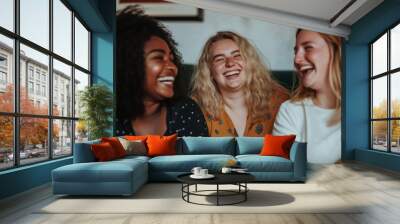 Three women are laughing and smiling at the camera. They are wearing different colored shirts and are sitting on a couch Wall mural