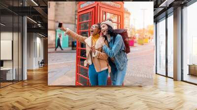 two friend, girlfriend and women using a mobile phone, camera and taking selfie against a red phonebox in the city of England.Travel Lifestyle concept Wall mural