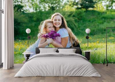 Two happy little girls sisters laughing, smiling, hugging, giving a kiss, holding flowers, sitting on green grass at summer meadow outdoor nature. Friendship and care concept. Wall mural