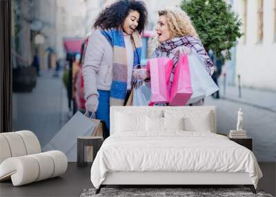 Two young multiracial female coworkers friends relieve stress after good work. Two different young curly woman relaxing with shoppig bags, wakls on street in winter time. Wall mural