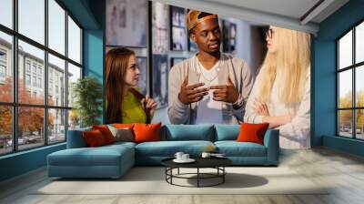 Young black hipster man talking and gesticulating with two girl friends in art photo gallery. Multi ethnic, shared values and culture friendship concept. Wall mural