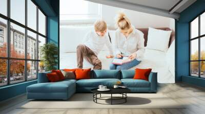 Young happy blond woman in blue jeans white shirt with little boy planing, writing on paper, sitting on sofa at living room interior. Wall mural