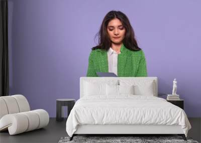 Young professional in a green coat reviews documents while holding a laptop against a soft purple background Wall mural