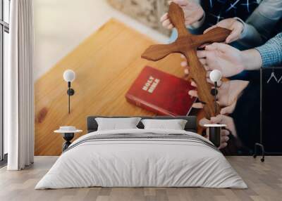 Close up of three Christian people hands holding wooden cross while praying together with the holy bible on wooden table indoor, Christian concept background with copy space Wall mural