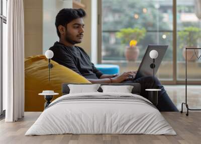 A indian man is sitting on a yellow bean bag chair and typing on a laptop Wall mural