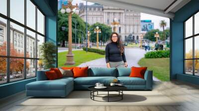 A young Latina woman poses in a park in Buenos Aires, Argentina, with historic architecture in the background. Wall mural