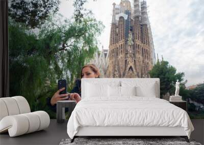 Positive young Latin woman traveler in warm outfit smiling and taking selfie on smartphone against Sagrada Familia by Antonio Gaudi during sightseeing tour in Barcelona, Spain Wall mural