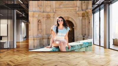 Young woman sitting at a historic site in Oaxaca, Mexico, wearing casual summer attire and sunglasses, smiling. Wall mural