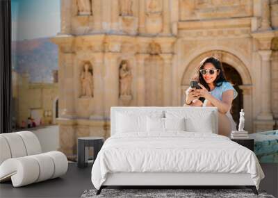 Young woman taking a selfie in front of a historic building in Oaxaca, Mexico, wearing casual summer attire. Wall mural