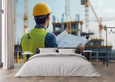 Construction worker reviewing blueprints on site, overseeing project development under a clear sky with cranes in the background. Wall mural