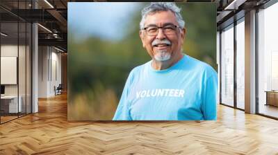 A cheerful older man with glasses wearing a blue volunteer t-shirt standing in front of a blurred natural background. Wall mural