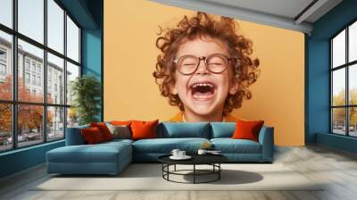 A joyful child with curly hair and glasses wearing a yellow sweater laughing heartily against a warm yellow background. Wall mural