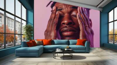 A man with a beard and dreadlocks wearing a ring with his eyes closed and hands on his face against a pink background. Wall mural