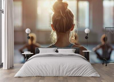 A woman with blonde hair in a bun wearing a gray tank top leading a yoga class with her back to the camera in a room with natural light coming through windows. Wall mural