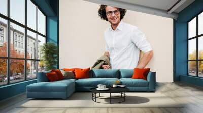 A young businessman in eyewear smiling broadly posing outdoors. Male entrepreneur resting in the city street. The curly guy in casual wears spectacles with curly hair walking outside after work Wall mural