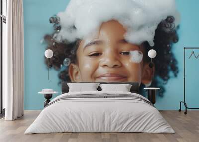 A young girl with curly hair smiling at the camera with soap bubbles on her face and hair set against a blue background. Wall mural