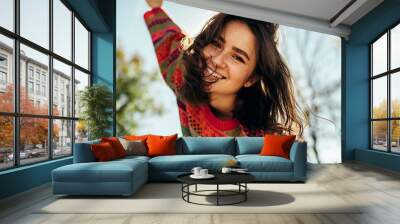 Bottom view close-up portrait of happy young woman smiling broadly with windy hair and freckles has joyful expression, wearing colorful knitted sweater with arms wide open, posing on nature sunlight Wall mural