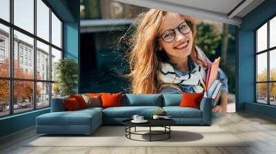 Candid portrait of a happy young student woman with long red hair smiling and wearing transparent eyeglasses standing next to the unversity and carrying lots of books and folders on a sunny day. Wall mural