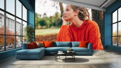 Caucasian young woman resting in the park. Female wearing an orange knitted sweater has pensive expression posing on the nature background. Wall mural