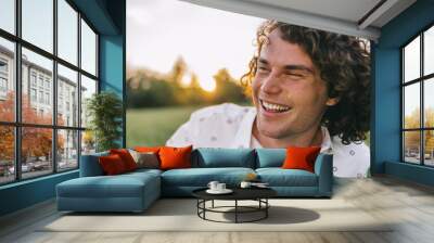 Close up portrait of happy young male with curly hair, smiling and relaxing on the green lawn in the park, listening funny story. Copy space for advertising. People and lifestyle concept. Wall mural