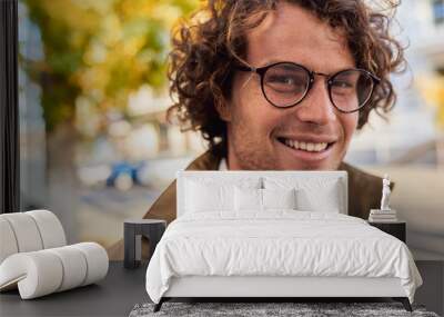 Closeup horizontal portrait of young happy business man with glasses smiling and posing outdoors. Male student in autumn street. Smart guy in casual wears spectacles with curly hair walking on street Wall mural