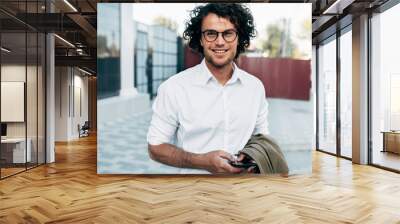 Confident businessman in eyewear smiling broadly posing outdoors. Male entrepreneur messaging on smartphone. Smart guy in casual wears spectacles with curly hair walking outside after work Wall mural