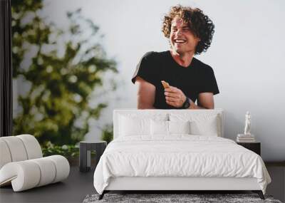 Confident happy guy with curly hair, smiling and wearing black t-shirt sitting on the street and preparing for exams. Freelancer businessman planning the day sitting on the city street Wall mural