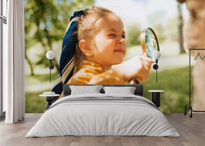 Curious kid with magnifying glass exploring the nature outdoor. Adorable little explorer girl playing in forest with magnifying glass. Child looking through magnifier on a sunny day in park Wall mural