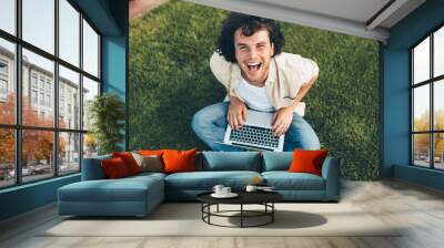 Happy man sitting on the ground in the park, working on laptop. Smiling man with curly hair using laptop for chatting online with friends, sitting on the grass outdoors. Technology, business, people Wall mural