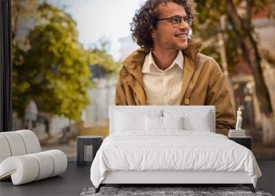 Happy young man with glasses reading and posing with book outdoors. College male student carrying books in campus in autumn street. Smiling guy wears spectacles and curly hair reading books outside Wall mural