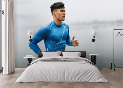 Horizontal outdoor image of young runner man running in the morning on the fog lake background. Fitness male exercising in the park and listenting the music on earphones. People and sport concept Wall mural