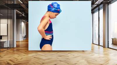 Horizontal side view of a cute little girl in goggles, and swimsuit, and a swimming cap looking down before swimming, isolated blue studio background. Wall mural