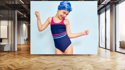 Horizontal studio image of a happy little girl in a swimsuit, swim goggles and swim cap making gesture during she wins the swimming contest, isolated over blue studio background. Wall mural